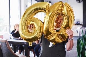 avec des ballons du numéro 60 dans les mains. femme âgée avec famille et amis célébrant un anniversaire à l'intérieur photo