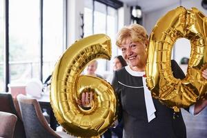 avec des ballons du numéro 60 dans les mains. femme âgée avec famille et amis célébrant un anniversaire à l'intérieur photo