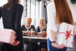 les jeunes filles tiennent des coffrets cadeaux derrière elles. femme âgée avec famille et amis célébrant un anniversaire à l'intérieur photo