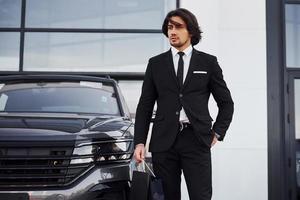 portrait d'un beau jeune homme d'affaires en costume noir et cravate à l'extérieur près d'une voiture moderne et avec des sacs à provisions photo