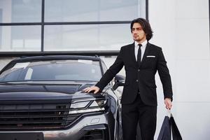 portrait d'un beau jeune homme d'affaires en costume noir et cravate à l'extérieur près d'une voiture moderne et avec des sacs à provisions photo