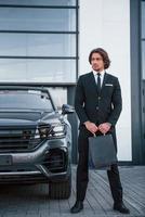 portrait d'un beau jeune homme d'affaires en costume noir et cravate à l'extérieur près d'une voiture moderne et avec des sacs à provisions photo