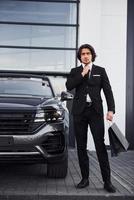 portrait d'un beau jeune homme d'affaires en costume noir et cravate à l'extérieur près d'une voiture moderne et avec des sacs à provisions photo