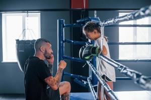 conception de l'aide. jeune entraîneur de boxe tatoué enseigne une jolie petite fille dans la salle de gym photo