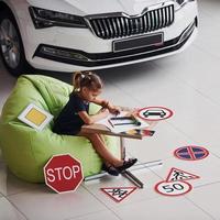 jolie petite fille est assise sur la chaise verte douce près de la table avec un crayon et des feuilles de papier. près de l'automobile moderne et des panneaux routiers sur le sol photo