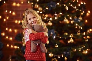 jolie petite fille en pull festif rouge avec ours en peluche à l'intérieur pour célébrer le nouvel an et les vacances de noël photo