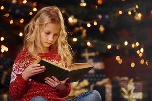 jolie petite fille en chandail festif rouge lisant un livre à l'intérieur pendant les vacances de noël photo