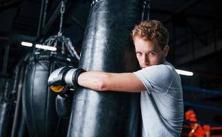 jeune boxeur fatigué en chemise blanche et avec des gants de protection s'appuyant sur un sac de boxe dans la salle de sport photo