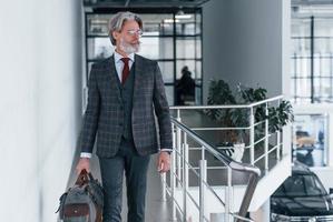 homme d'affaires senior en costume et cravate avec cheveux gris et barbe marchant à l'intérieur avec sac photo