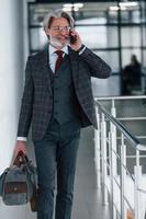 homme d'affaires senior en costume et cravate avec cheveux gris et barbe marchant à l'intérieur avec sac photo
