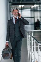 homme d'affaires senior en costume et cravate avec cheveux gris et barbe marchant à l'intérieur avec sac photo