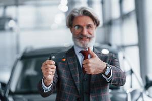 homme aux cheveux gris et à la moustache debout contre une voiture moderne à l'intérieur avec une carte de crédit en main photo