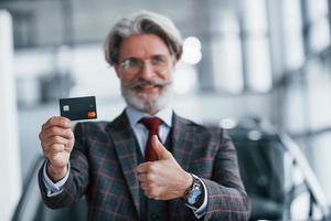homme aux cheveux gris et à la moustache debout contre une voiture moderne à l'intérieur avec une carte de crédit en main photo
