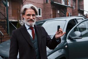homme âgé à la mode avec des cheveux gris et une barbe debout à l'extérieur dans la rue près de sa voiture avec les clés en main photo