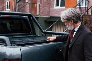 un homme âgé à la mode aux cheveux gris et à la barbe ferme le coffre de sa voiture photo