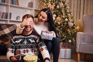 petite amie couvre les yeux de son mari et donne la surprise du nouvel an dans la chambre décorée de noël photo