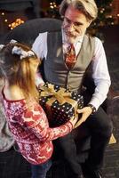 homme âgé à la mode joyeuse avec des cheveux gris et une barbe assis avec une petite fille dans la salle de noël décorée photo