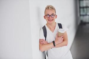 étudiant en vêtements décontractés et avec sac à dos se tient à l'intérieur contre un mur blanc avec une tasse de boisson photo