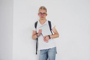 étudiant en vêtements décontractés et avec sac à dos se tient à l'intérieur contre un mur blanc avec un ordinateur portable photo