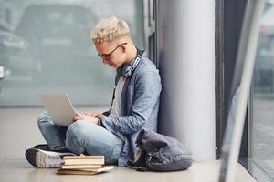 jeune homme hipster assis à l'intérieur près d'un fond gris avec un ordinateur portable et des livres photo