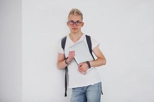 étudiant en vêtements décontractés et avec sac à dos se tient à l'intérieur contre un mur blanc avec un ordinateur portable photo