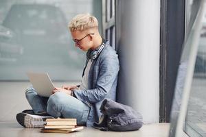 jeune homme hipster assis à l'intérieur près d'un fond gris avec un ordinateur portable et des livres photo