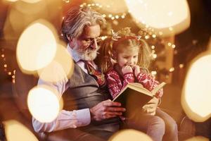 homme âgé à la mode joyeuse avec des cheveux gris et une barbe assis avec une petite fille dans un livre de lecture de salle de noël décoré photo