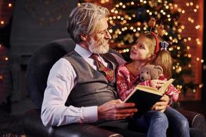 homme âgé à la mode joyeuse avec des cheveux gris et une barbe assis avec une petite fille dans un livre de lecture de salle de noël décoré photo