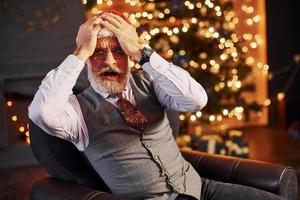 portrait d'un senior élégant aux cheveux gris et à la barbe assis dans une pièce décorée et au chapeau de noël photo