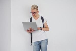 étudiant en vêtements décontractés et avec sac à dos se tient à l'intérieur contre un mur blanc avec un ordinateur portable photo