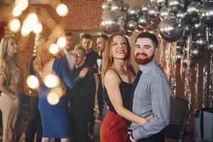 jeune homme barbu avec sa petite amie debout ensemble contre leurs amis dans une salle décorée de noël et célébrant le nouvel an photo