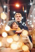 jeune couple positif dans des vêtements chauds s'embrassant sur la rue décorée de noël photo