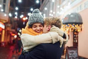 jeune couple positif dans des vêtements chauds s'embrassant sur la rue décorée de noël photo