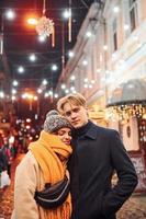 jeune couple positif dans des vêtements chauds s'embrassant sur la rue décorée de noël photo