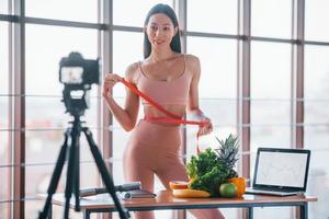 jeune vlogger de fitness faisant de la vidéo à l'intérieur en se tenant près de la table avec de la nourriture saine et un ordinateur portable photo