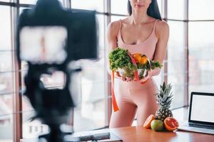 jeune vlogger de fitness faisant de la vidéo à l'intérieur en se tenant près de la table avec de la nourriture saine et un ordinateur portable photo