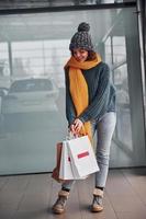 belle fille gaie en écharpe jaune et en vêtements chauds debout à l'intérieur avec des sacs à provisions dans les mains photo