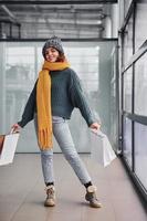 belle fille gaie en écharpe jaune et en vêtements chauds debout à l'intérieur avec des sacs à provisions dans les mains photo