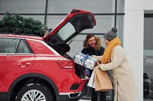 jeune couple avec des coffrets cadeaux est près de la voiture avec un arbre sur le dessus. ensemble à l'extérieur en hiver photo