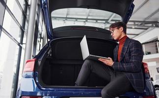 gars en tenue de soirée et en lunettes assis à l'arrière de la voiture avec un ordinateur portable dans les mains photo