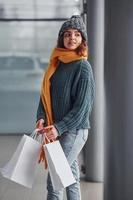 belle fille gaie en écharpe jaune et en vêtements chauds debout à l'intérieur avec des sacs à provisions dans les mains photo