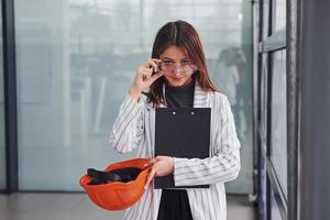 portrait de jeune travailleuse avec casque de protection, lunettes et bloc-notes. debout au bureau photo