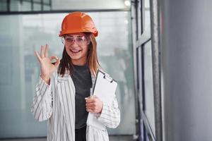 portrait de jeune travailleuse avec casque de protection, lunettes et bloc-notes. debout au bureau photo