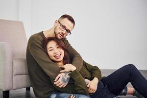 heureux couple multiethnique dans des vêtements décontractés assis ensemble à l'intérieur dans le studio. mec caucasien avec petite amie asiatique photo