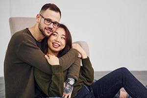 heureux couple multiethnique dans des vêtements décontractés assis ensemble à l'intérieur dans le studio. mec caucasien avec petite amie asiatique photo