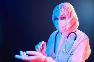 infirmière en masque et uniforme blanc et avec stéthoscope debout dans une pièce éclairée au néon photo