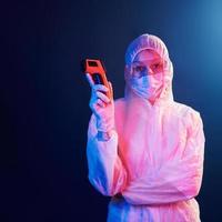 infirmière en masque et uniforme blanc debout dans une pièce éclairée au néon et tenant un thermomètre infrarouge. arrêter la propagation du coronavirus photo