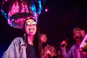 jeune fille tenant des pilules de drogue sur togue devant des jeunes qui s'amusent dans une boîte de nuit avec des lumières laser colorées photo