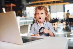 enfant intelligent dans des vêtements décontractés avec un ordinateur portable sur la table s'amuser avec une loupe photo