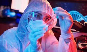 infirmière en masque et uniforme blanc, tenant un tube avec du liquide et assise dans un laboratoire éclairé au néon avec ordinateur et équipement médical à la recherche d'un vaccin contre le coronavirus photo